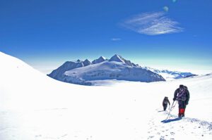 Two people walking in the snow away from a mountain. There is a blue sky with a single cloud.