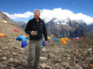 Dave Smith in Bhutan standing in front of a mountain with tents pitched in the background.