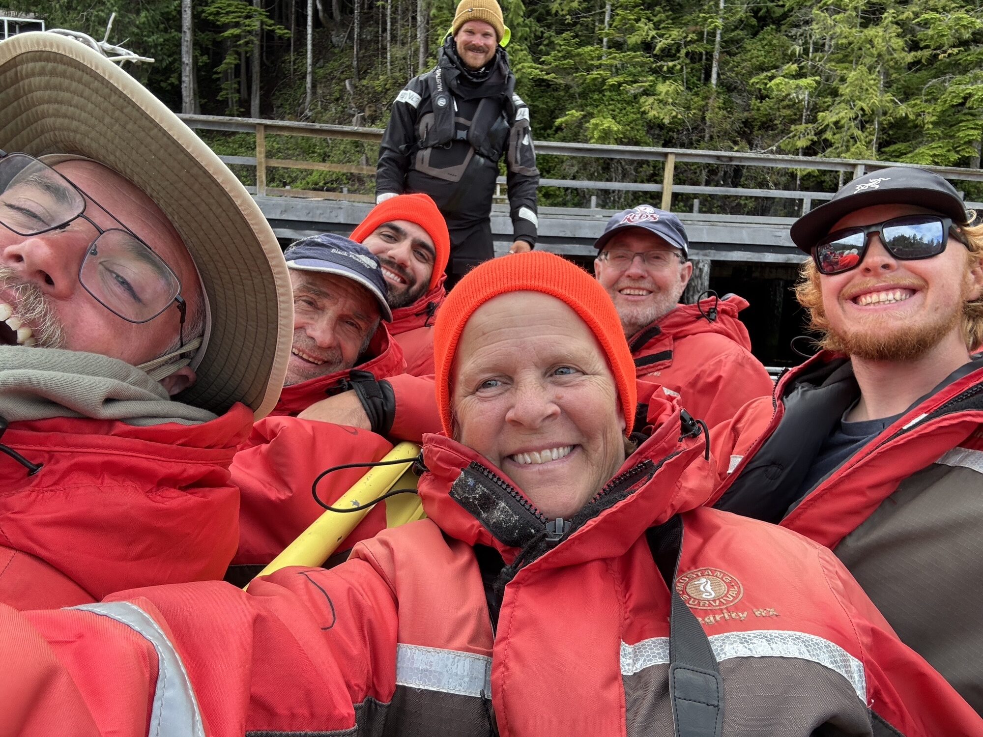 Group of travelers in red flotation suits gather for a selfie on a dock, with a forested shoreline and another person standing in the background.