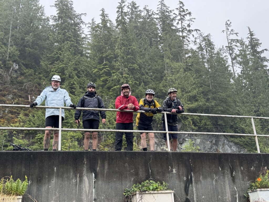 Five cyclists in rain gear and helmets stand along a railing overlooking a forested landscape on an overcast day.