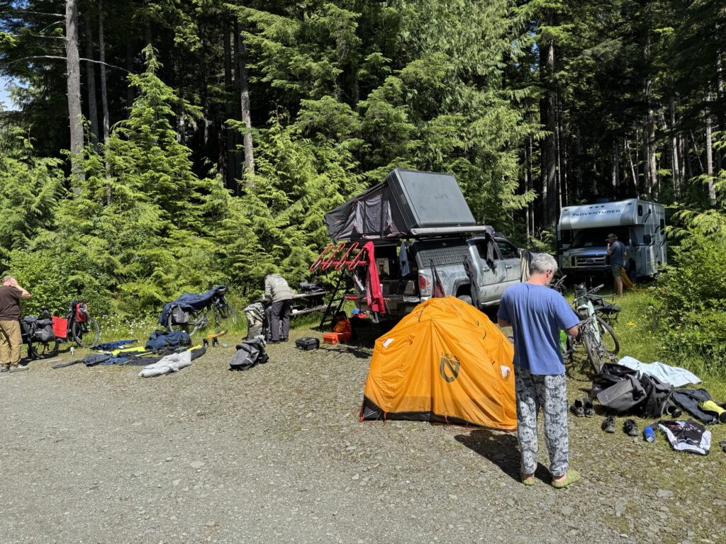 Cyclists and campers organize gear at a forest campsite featuring an orange tent, bicycles, a camper truck with a rooftop tent, and an RV.