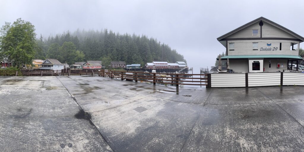 Rain-slicked waterfront boardwalk and marina beside a building overlooking a foggy harbour, with colorful shoreline buildings and forested hills in the background.