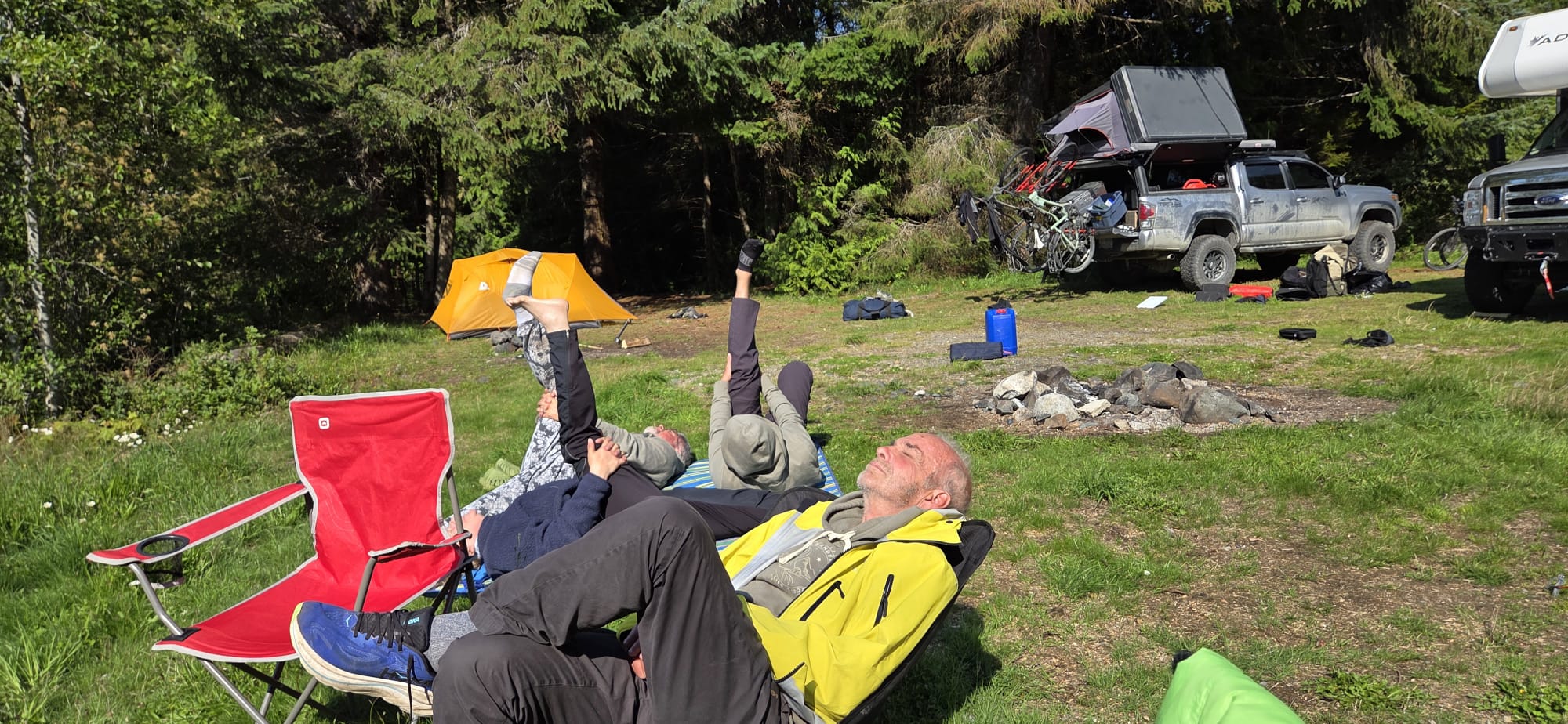 People relaxing and stretching at a forested lakeside campsite with camping chairs, an orange tent, a rooftop tent, and bicycles mounted on a pickup truck.