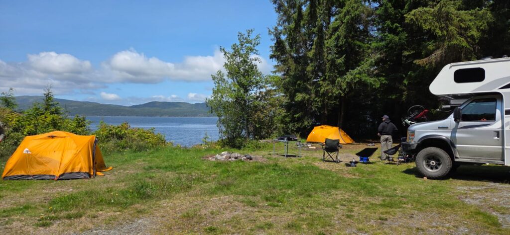 Lakeside campground with two orange tents, camping chairs, an RV, and mountain views across the water under a blue sky.
