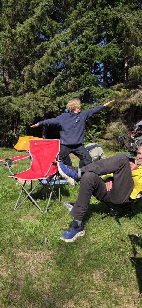 Camper practicing a standing stretch beside a red camp chair at a forested lakeside campsite, with tents and camping gear visible in the background.