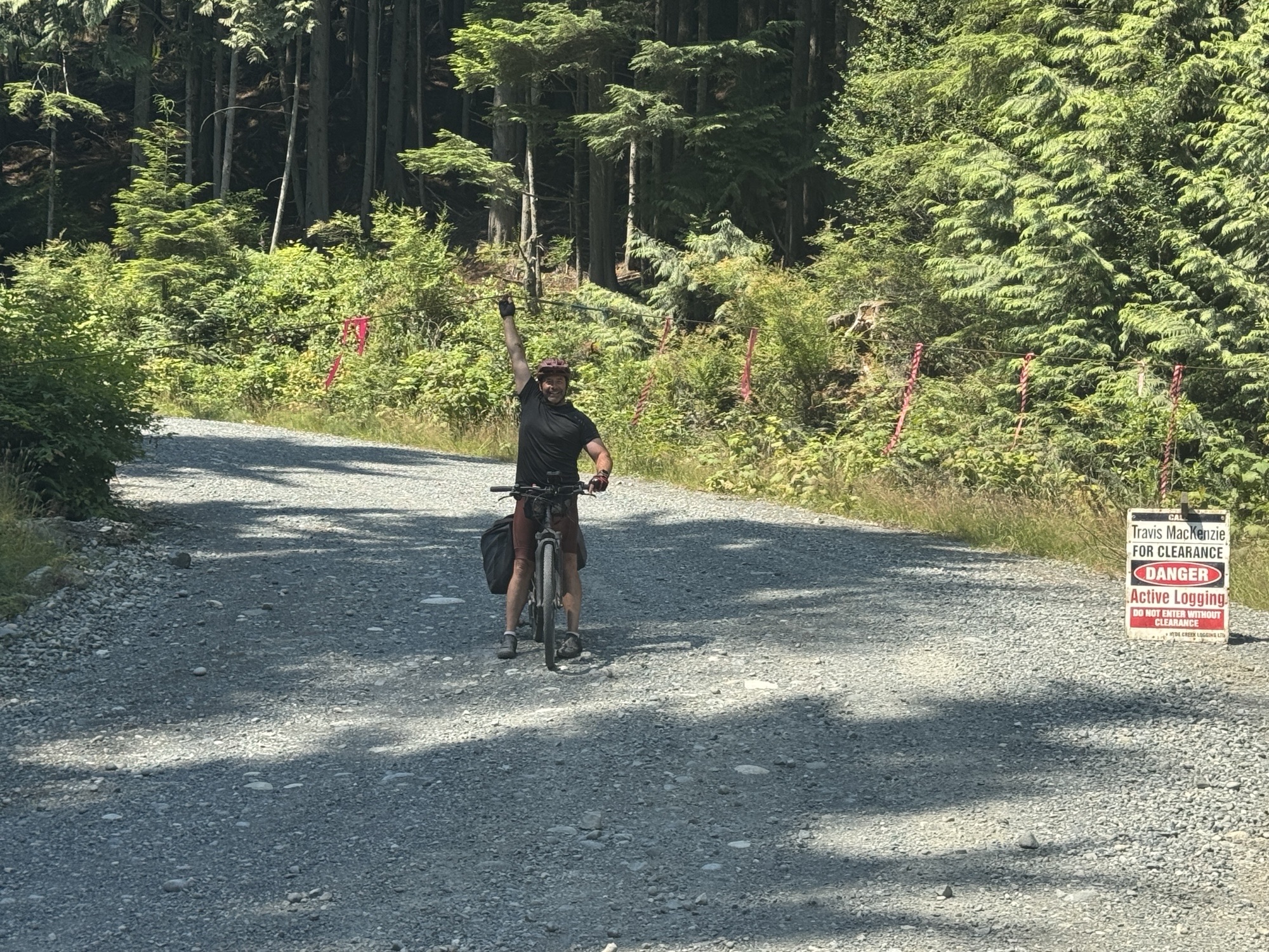 Cyclist on a gravel forest road raising one arm while riding through a wooded area, with logging activity warning signs visible along the roadside.
