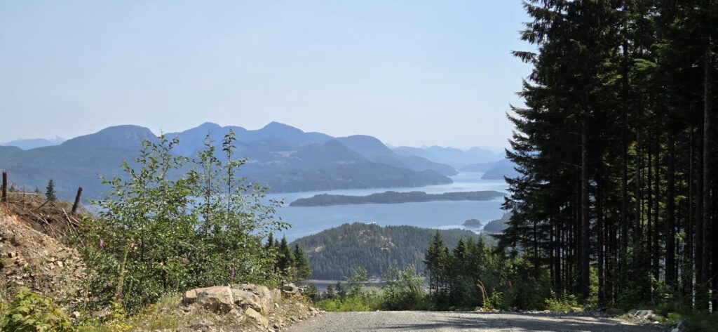 Scenic mountain and coastal landscape viewed from a forest road, overlooking forested islands, blue water, and layered mountain ranges in the distance.
