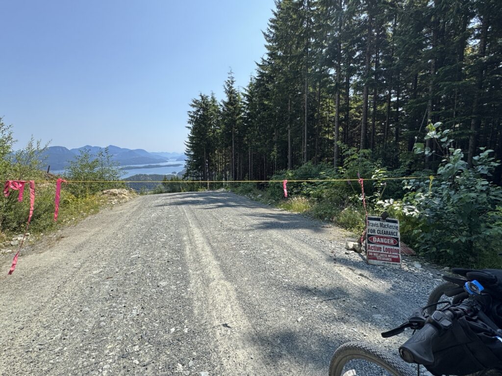 Gravel logging road blocked by a rope barrier, with an active logging warning sign beside the road and mountain and coastal views visible in the distance.