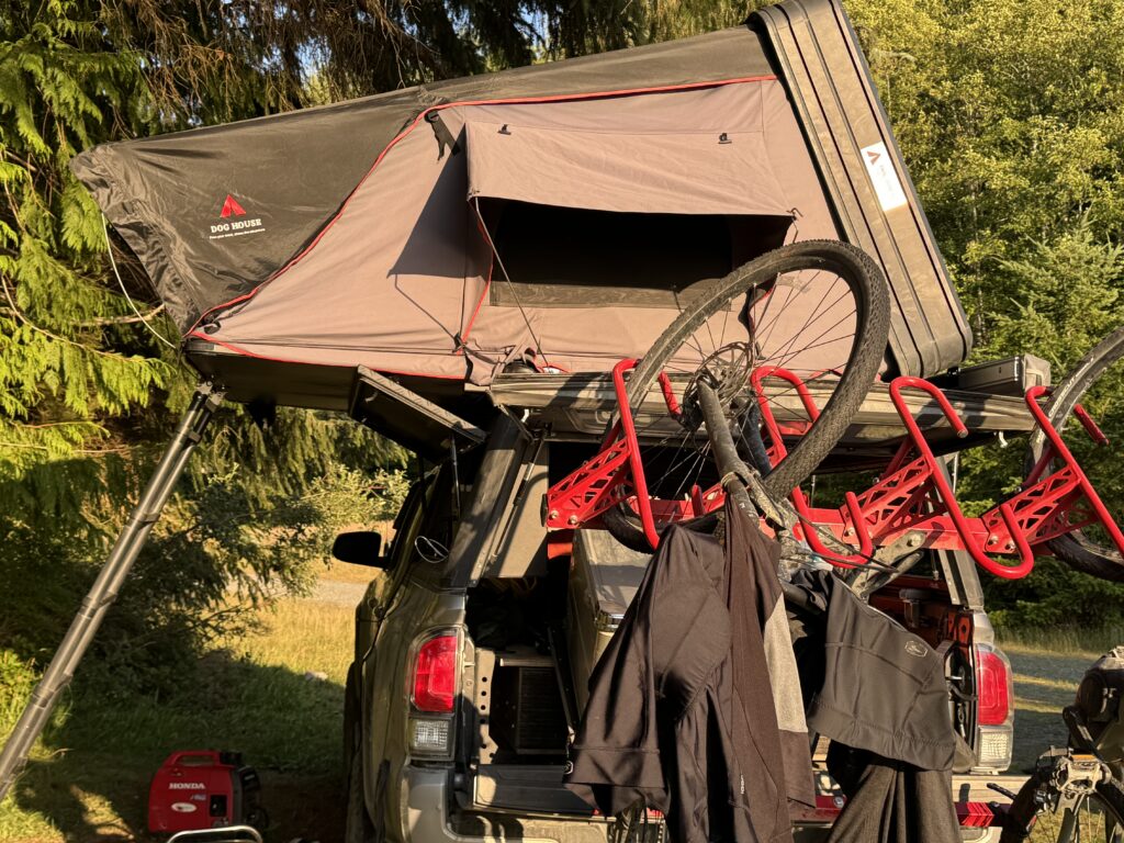 Rooftop tent mounted on a pickup truck at a forest campsite, with mountain bikes secured on a rear rack and camping gear visible below.
