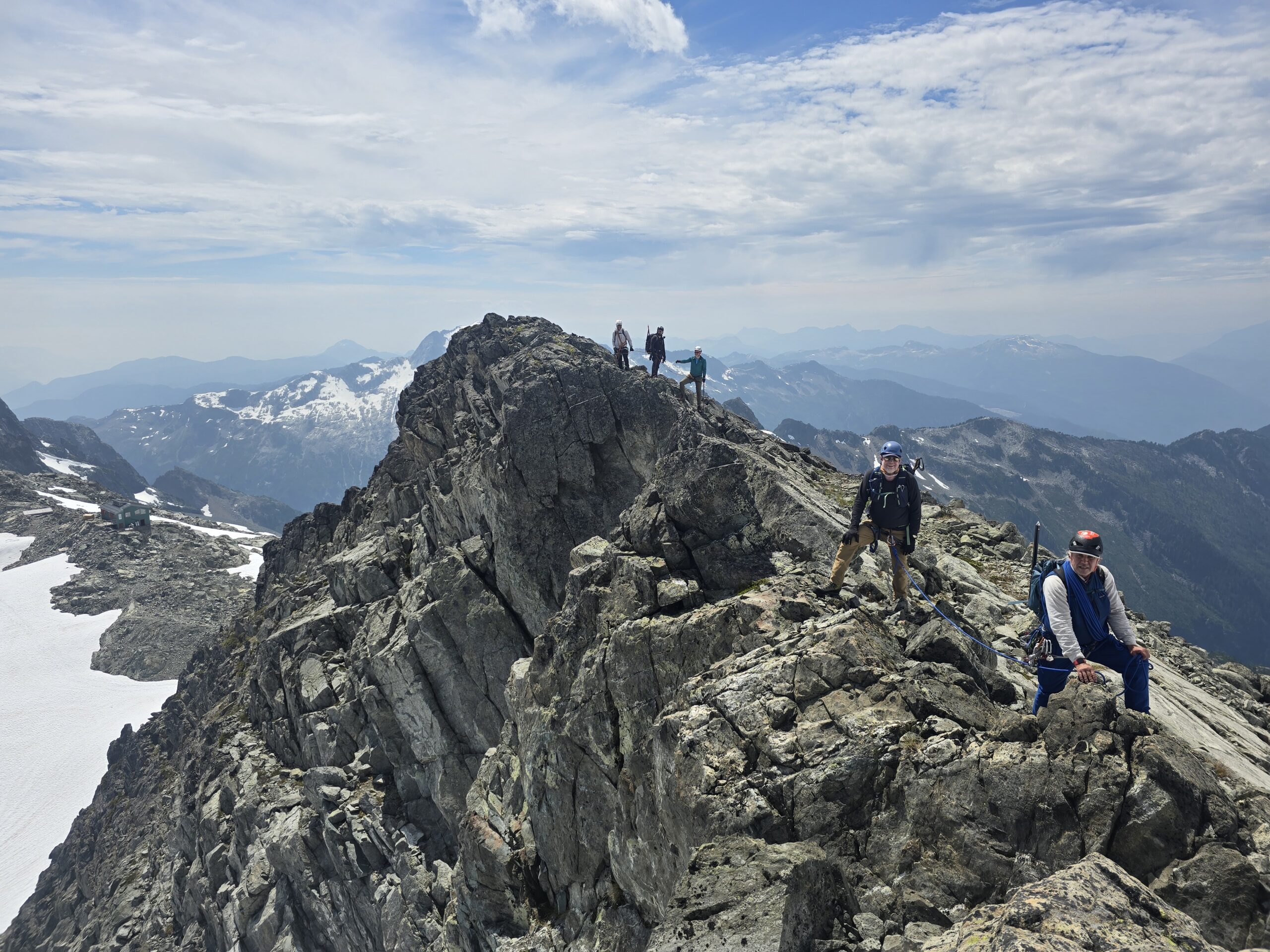 A group of climbers wearing helmets and safety gear stands along a rugged rocky mountain ridge, with snow-covered slopes and distant mountain ranges beneath a partly cloudy sky.
