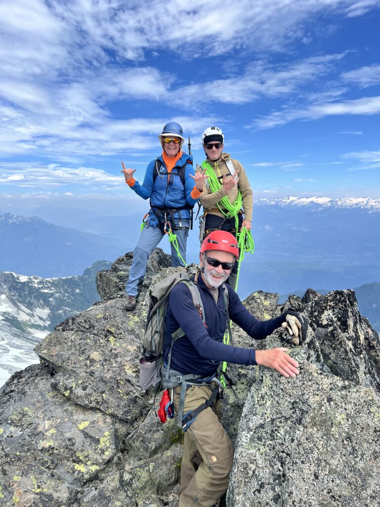 Three climbers in helmets and harnesses pose on a rocky mountain summit, with climbing ropes, alpine peaks, and a bright blue sky with scattered clouds in the background.