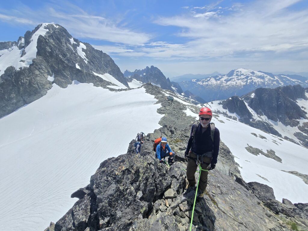 Climbers traverse a narrow rocky ridge above a snow-covered alpine landscape, with one climber in the foreground and glacier-covered mountains stretching into the distance.