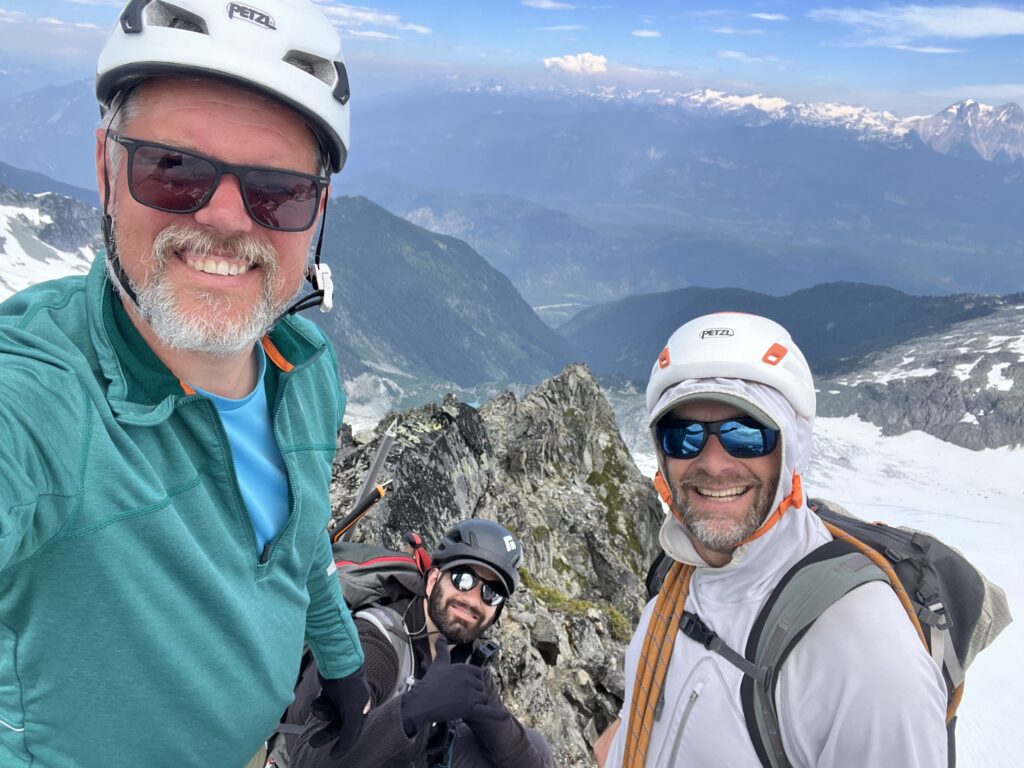 Three helmeted mountaineers pause for a photo on a rocky alpine ridge, with snowfields, rugged peaks, and a sweeping mountain valley visible in the background under a clear blue sky.