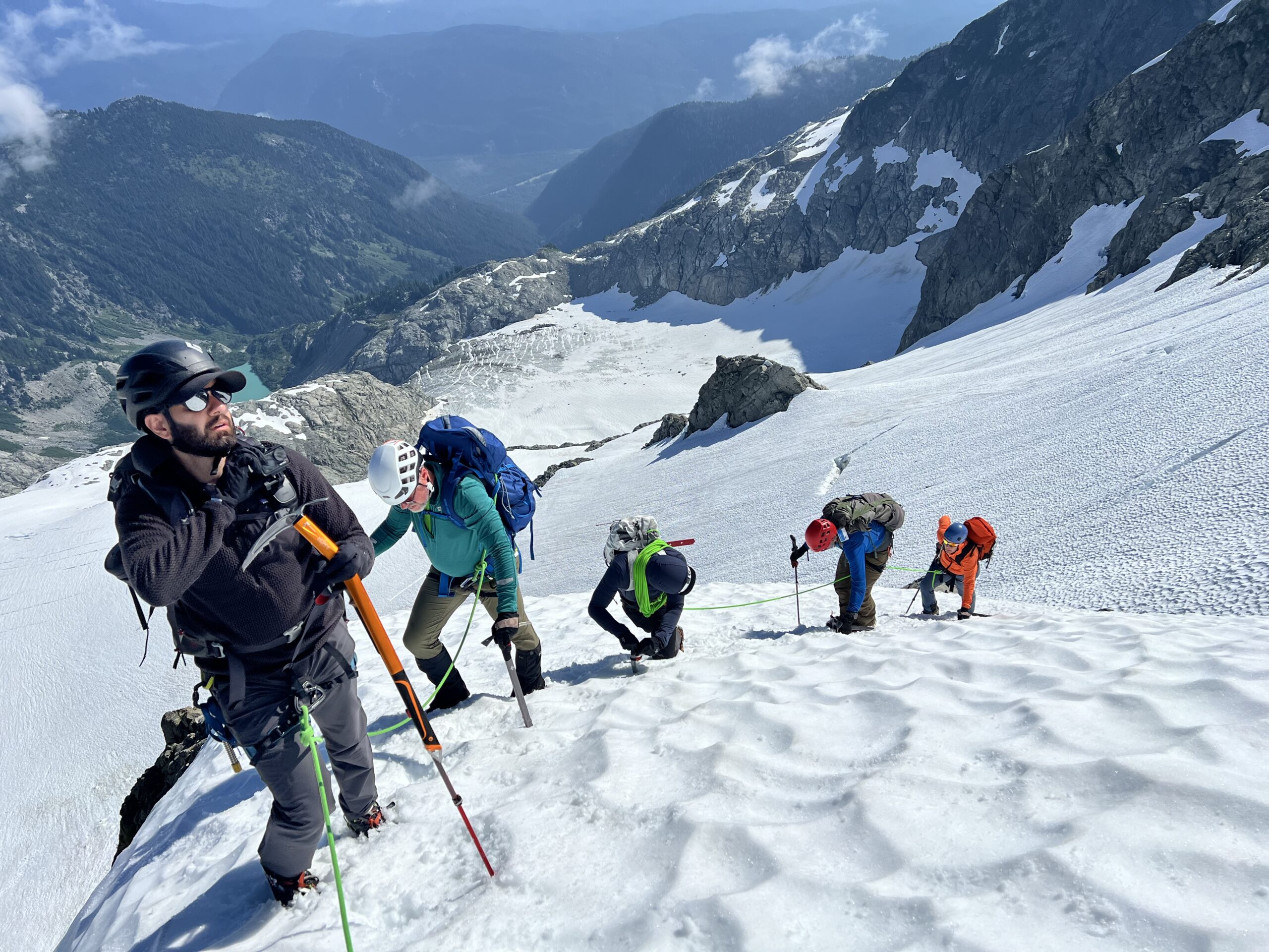 Roped team of mountaineers using ice axes to climb a steep snow-covered glacier above a rugged alpine valley and rocky mountain peaks.