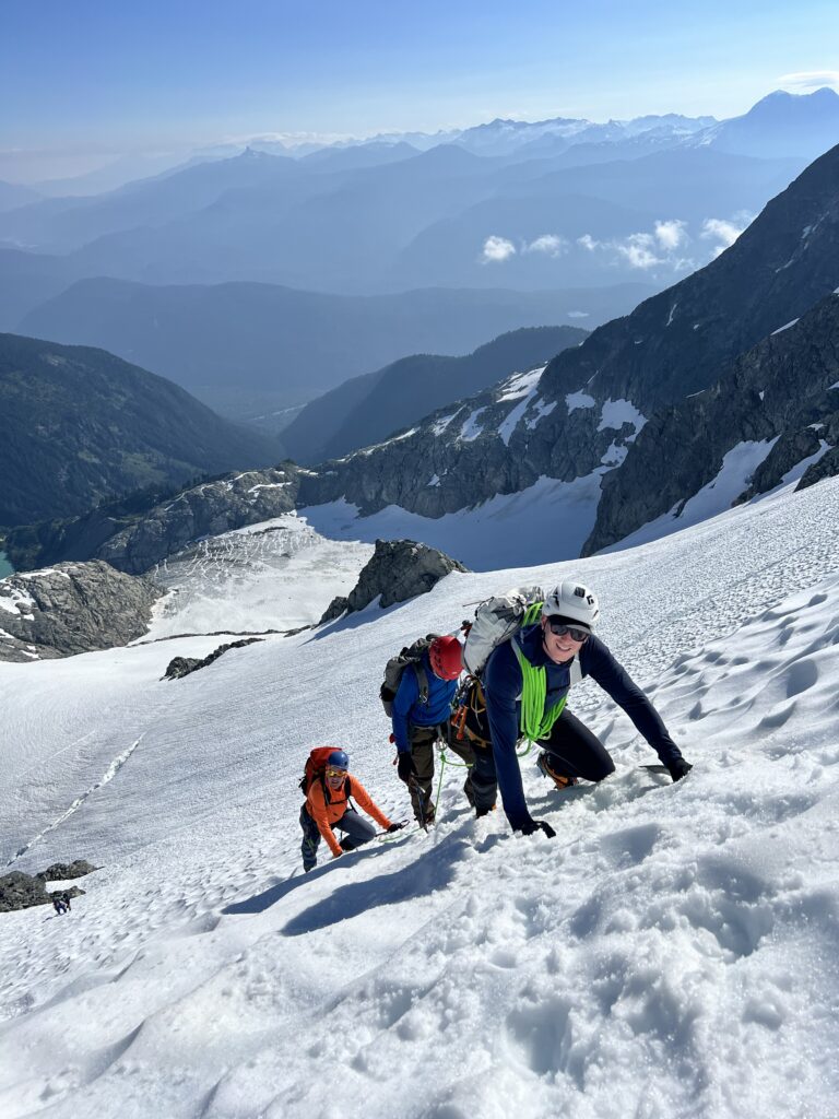 Three climbers ascending a steep snowy slope while roped together, with expansive mountain ranges and glaciers stretching into the distance.