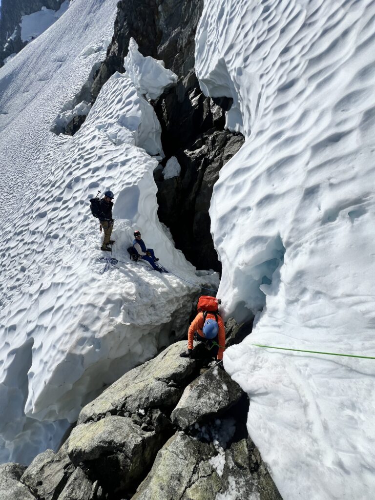 Mountaineers navigating a narrow passage between towering snow and ice formations, crossing rocks beside a deep glacier crevasse.