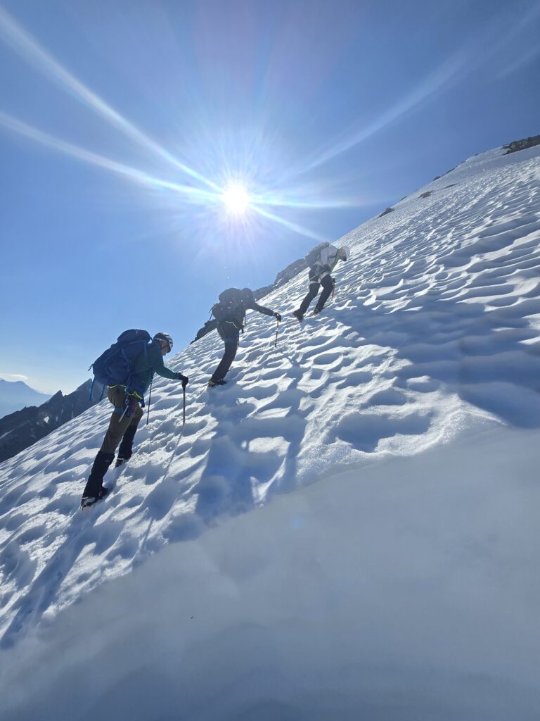 Three mountaineers use ice axes to climb a steep snow-covered slope beneath a bright sun and clear blue sky in alpine terrain.