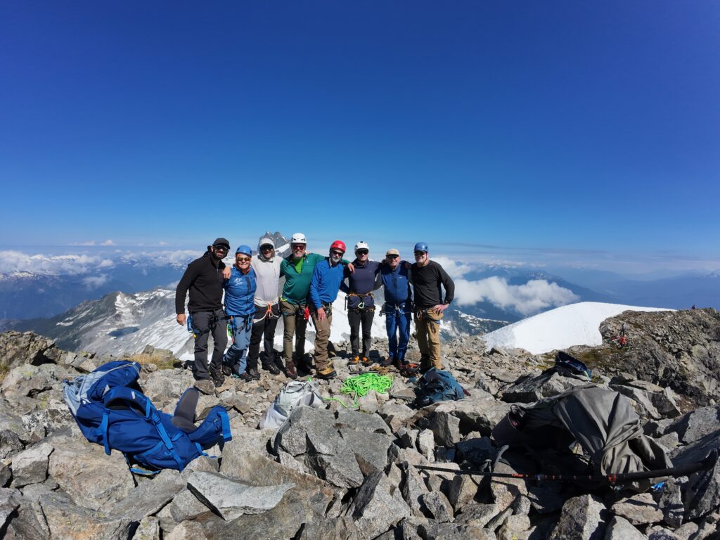 Group of eight climbers standing on a rocky mountain summit with snow-covered peaks and clouds stretching across the horizon behind them.
