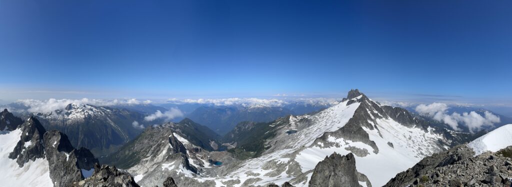 Panoramic view from a mountain summit showing rugged snow-covered ridges, glaciers, rocky peaks, and distant alpine valleys under a clear blue sky.