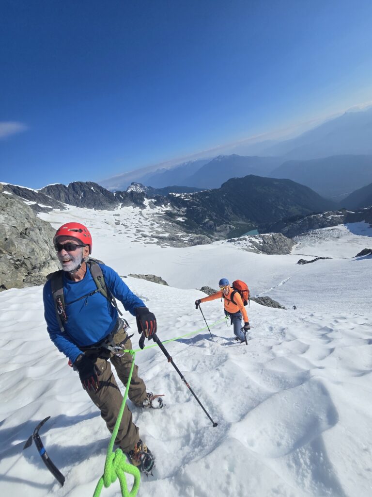 Roped mountaineers descending a steep glacier using ice axes and crampons, with expansive snowfields, rocky ridges, and alpine lakes visible below.