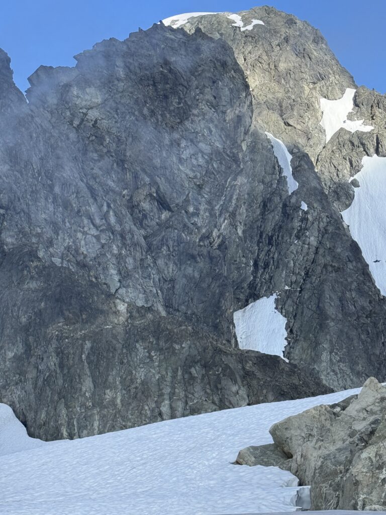 Towering rocky mountain face rising above a snow-covered glacier, with patches of lingering snow beneath a clear blue sky.