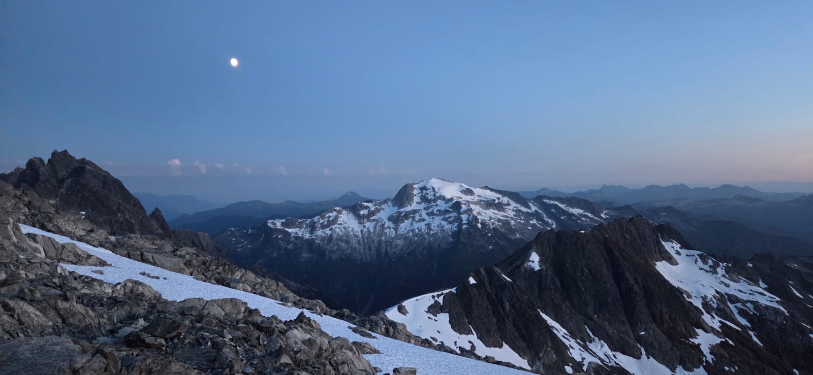 Panoramic view of rugged, snow-covered mountain peaks at twilight, with rocky ridges in the foreground and a pale moon visible in the sky.