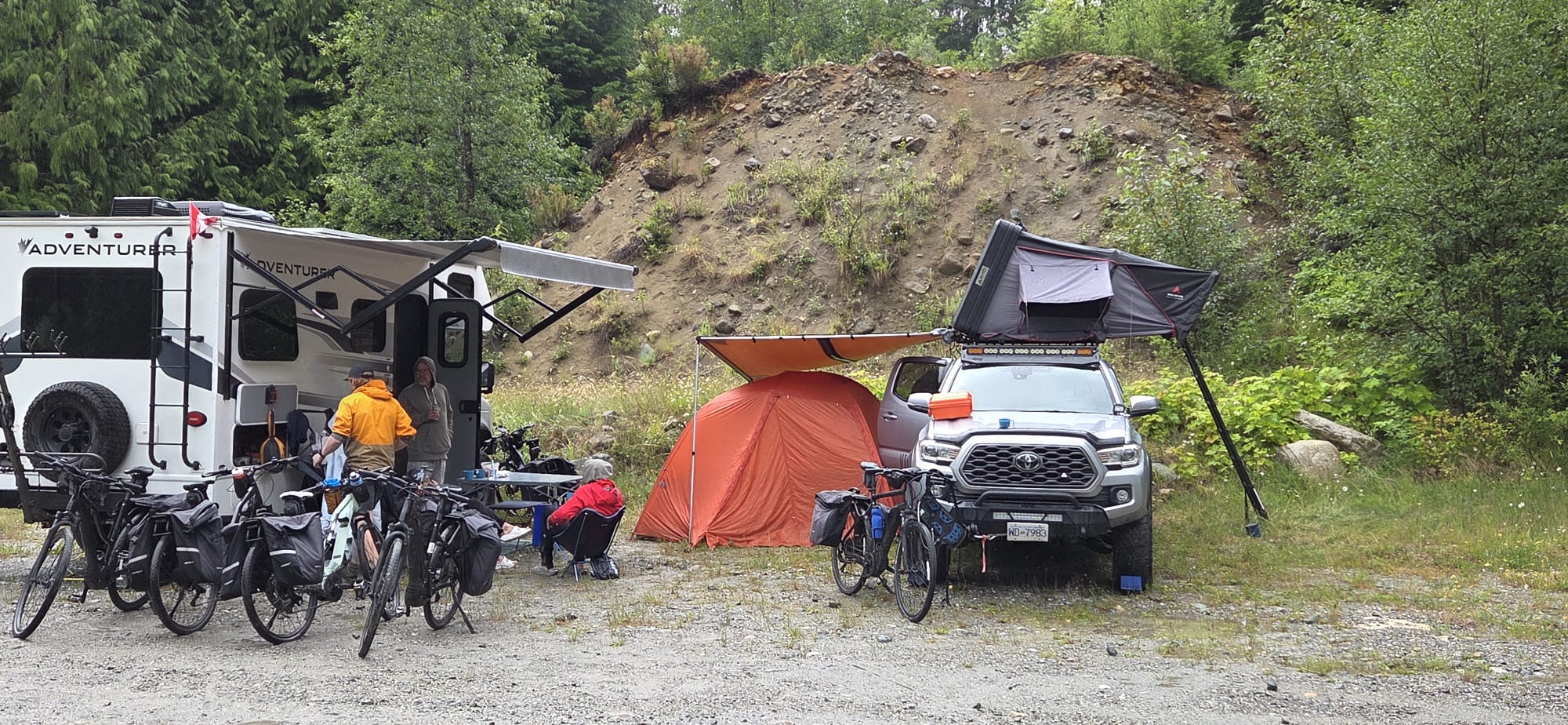 A group sets up camp with a camper van, a pickup truck with a rooftop tent, several bicycles, and outdoor gear near a wooded hillside.