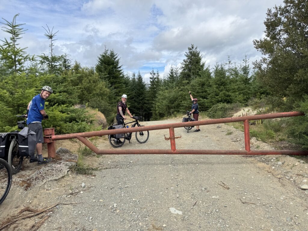 Three cyclists stop at a red metal gate blocking a gravel trail through a forested area under a partly cloudy sky.