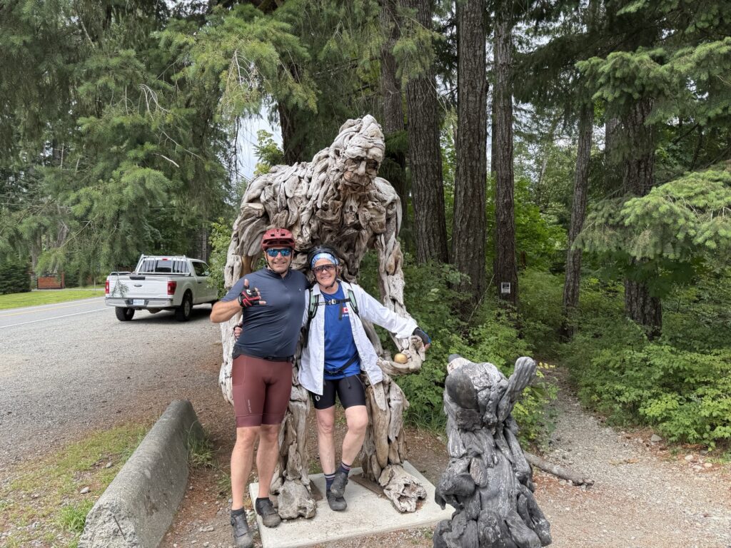 Two cyclists pose smiling in front of a large wooden Bigfoot sculpture in a forested area.