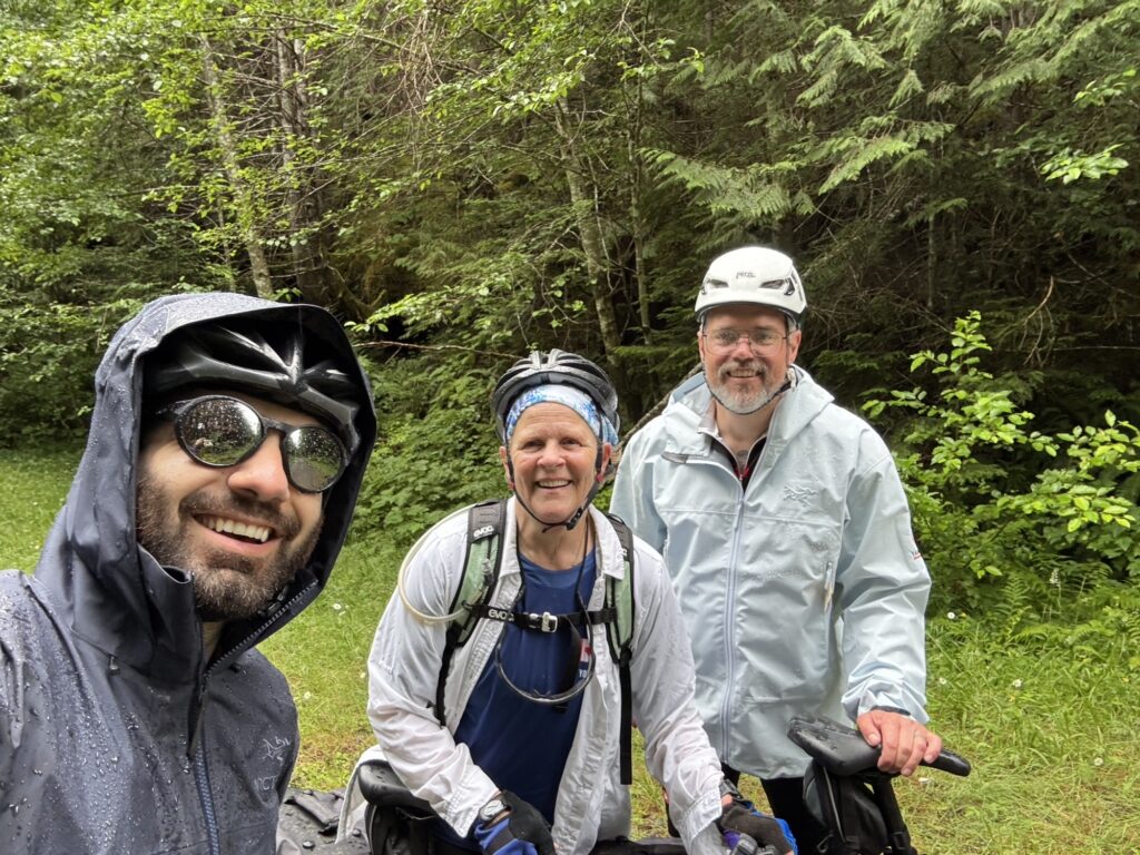 Three people wearing cycling helmets and rain jackets smile at the camera while standing with their bicycles in a forest clearing.
