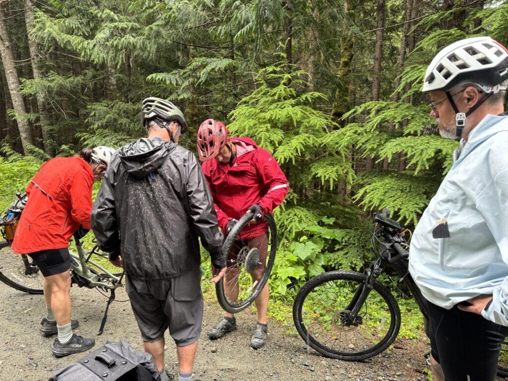 Several cyclists stop on a gravel road in a forest while one person repairs a bicycle wheel.