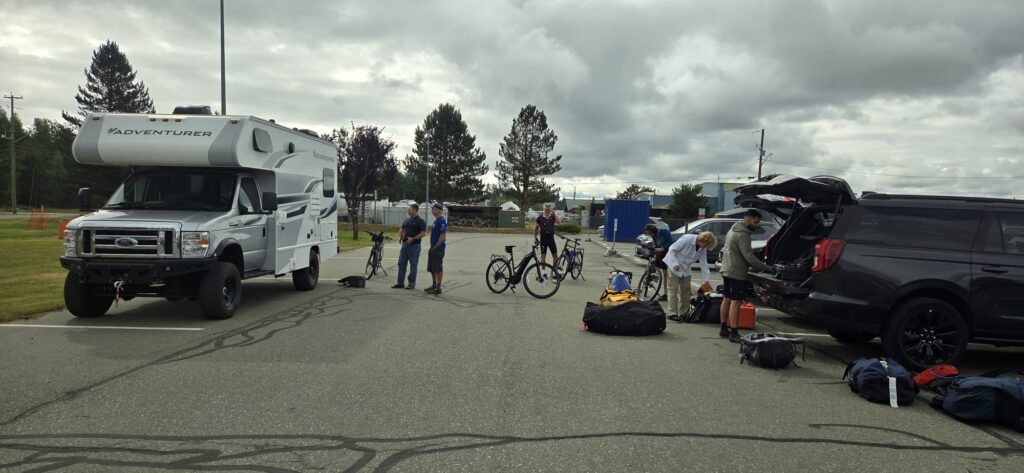 People unload bicycles and travel gear from vehicles in a parking lot beside a motorhome under a cloudy sky.