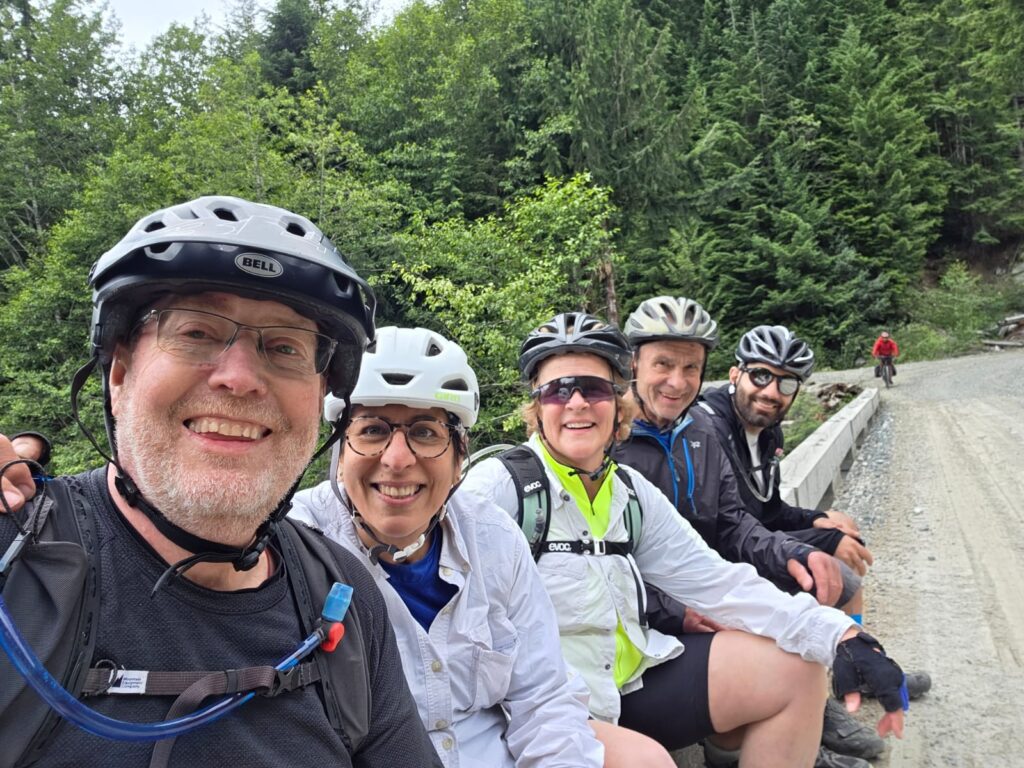 Five cyclists in helmets and outdoor gear pose happily on a forest trail with lush greenery.
