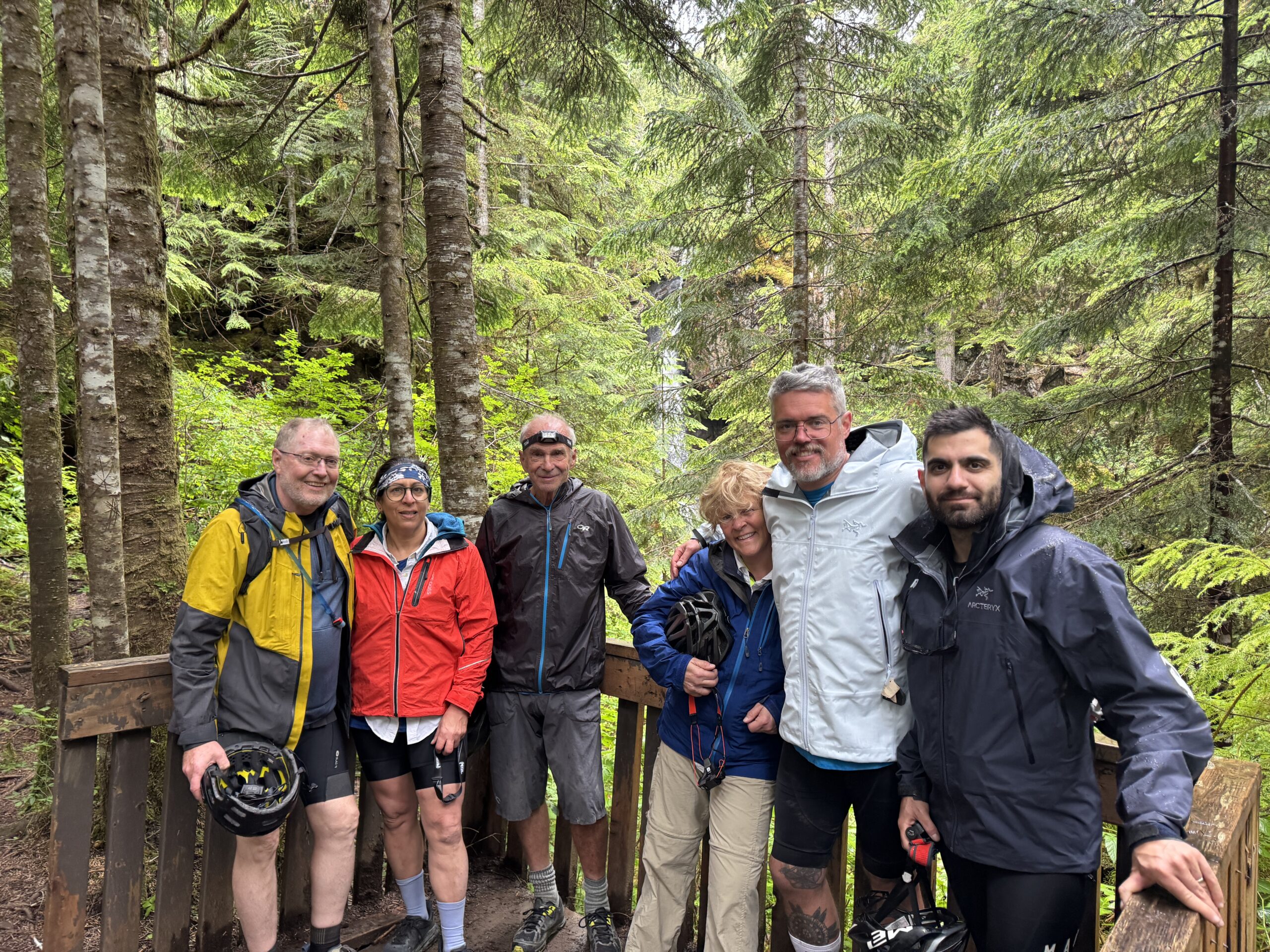 Six cyclists standing together on a wooden viewing platform in a dense evergreen forest, with a small waterfall visible in the background.