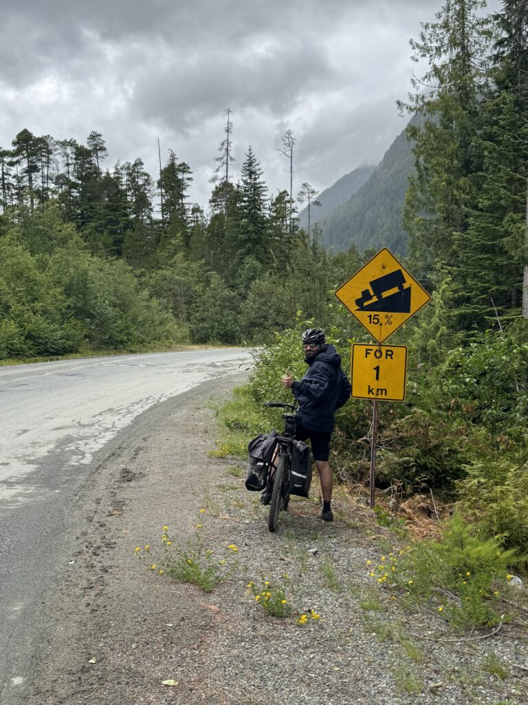 Cyclist with a loaded touring bicycle stopped beside a winding mountain road near a steep 15 percent grade warning sign, surrounded by forested slopes under cloudy skies.
