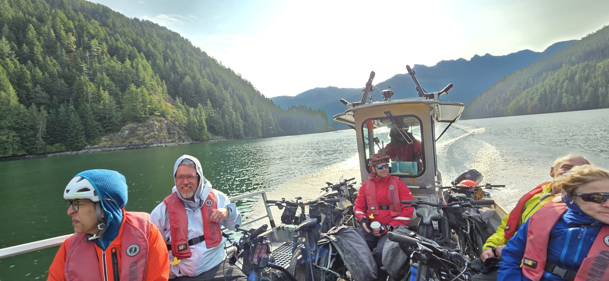 Passengers and loaded touring bicycles aboard a small boat crossing a forest-lined mountain lake, with calm water and scenic peaks in the background.