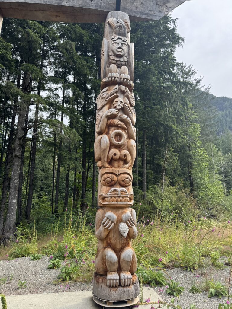 Tall wooden totem pole featuring carved figures and animal motifs, standing in a clearing surrounded by evergreen forest and wildflowers.