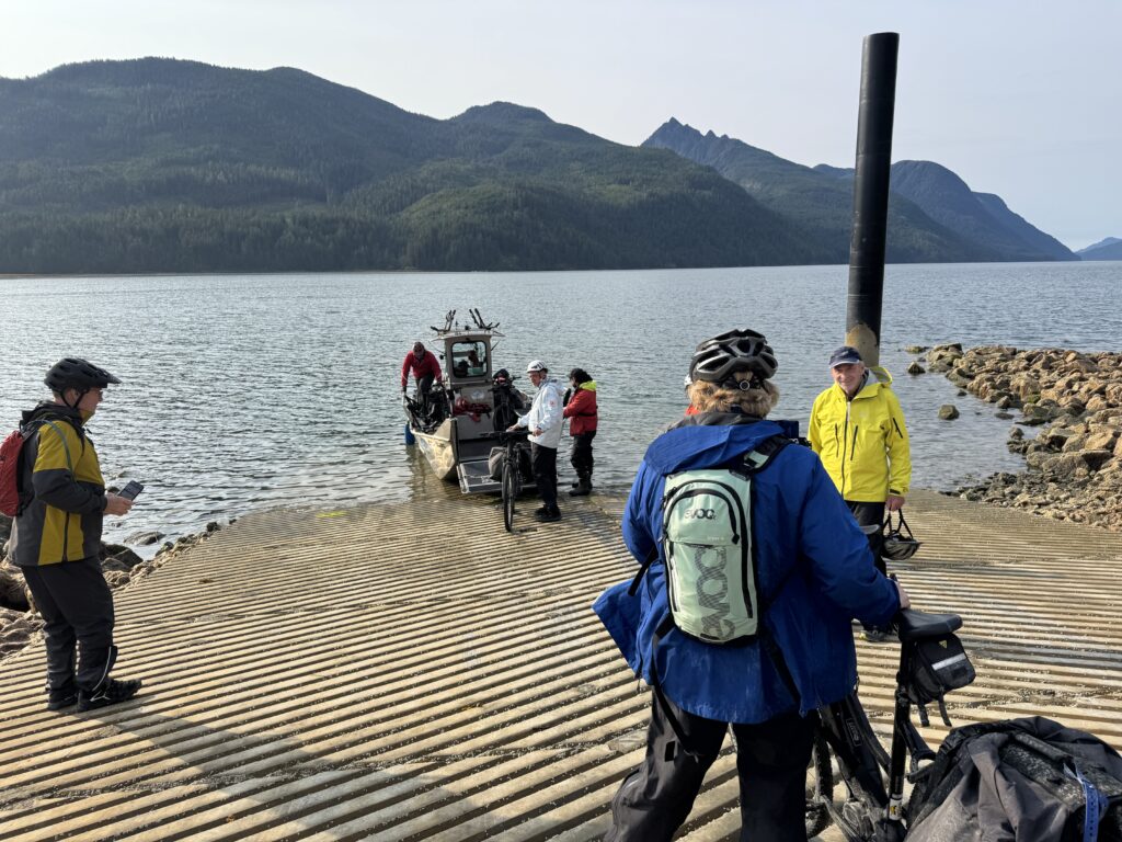 Cyclists gather on a lakeside boat ramp as a small ferry unloads passengers and bicycles, with forested mountains and calm water in the background.