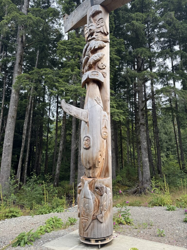 Carved wooden totem pole featuring animal and human figures, viewed from the side in a forest clearing surrounded by tall evergreen trees.