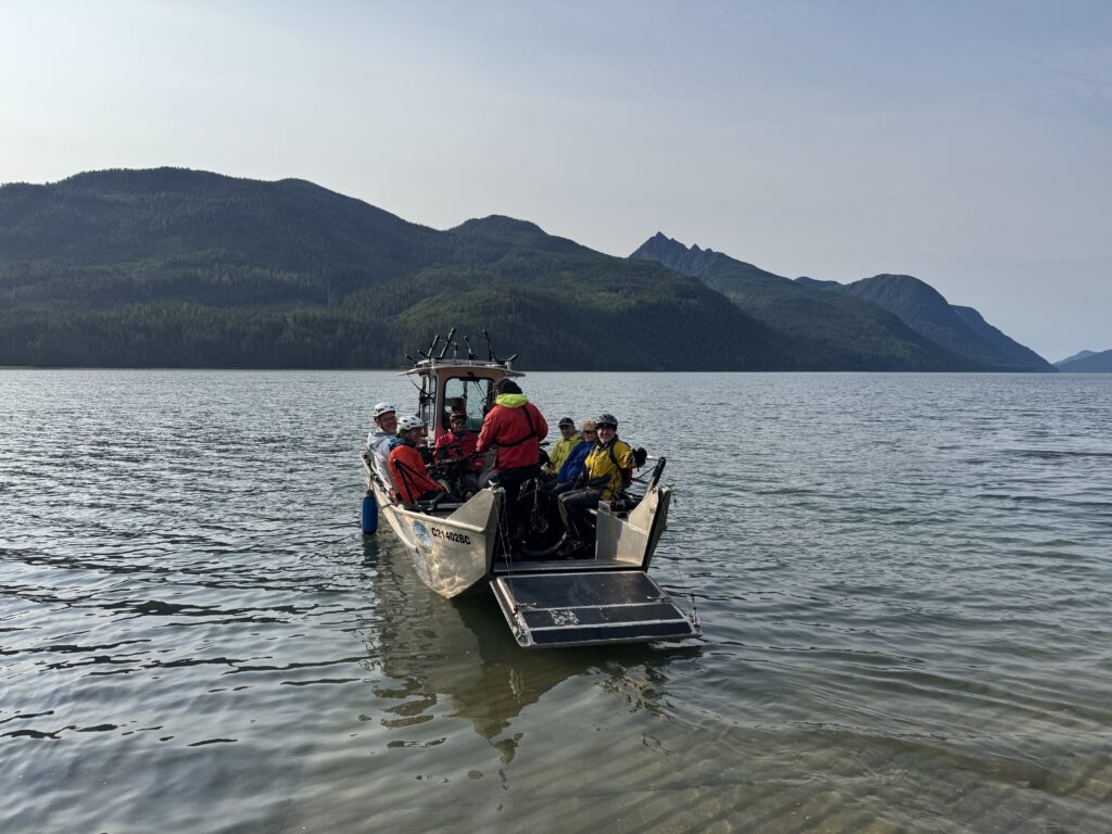 Small ferry carrying cyclists and bicycles across a mountain lake, with forest-covered peaks and clear water stretching into the distance.