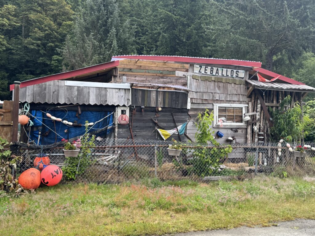 Rustic waterfront building in Zeballos made from weathered wood, decorated with fishing nets, buoys, and marine equipment, with a forested hillside in the background.