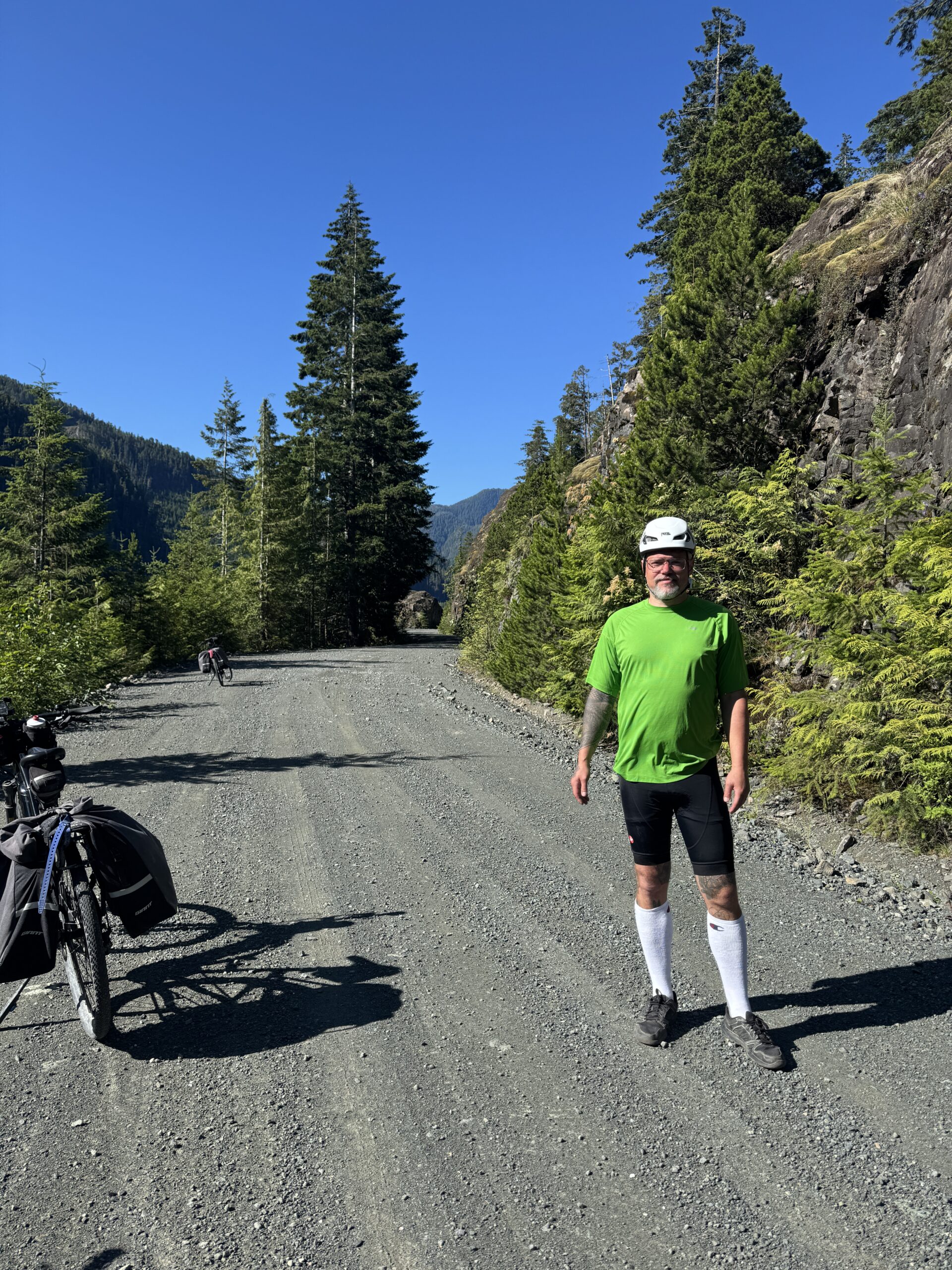 Cyclist standing beside a loaded touring bike on a gravel road surrounded by evergreen forest and rocky mountains under a clear blue sky.