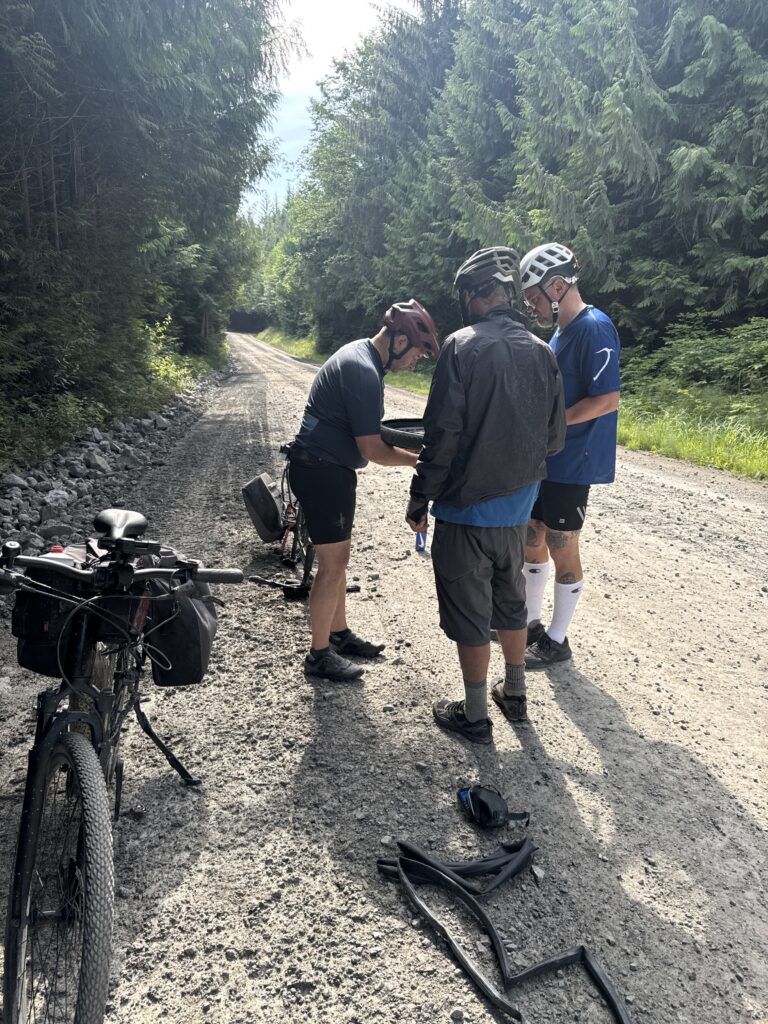 Three cyclists wearing helmets stop on a gravel forest road to repair a bicycle tire. A touring bicycle with panniers is parked nearby, and spare inner tubes and repair gear are laid out on the road beside tall evergreen trees.