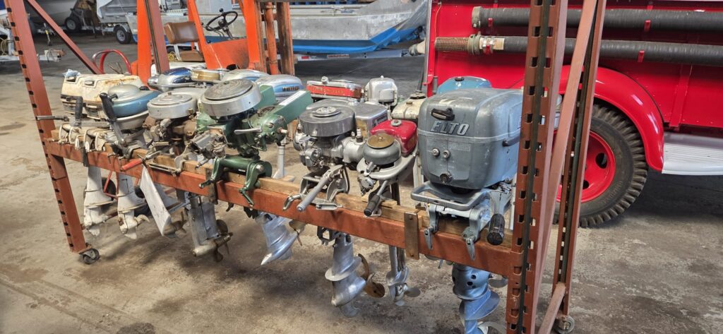 Collection of vintage outboard boat motors displayed on a rolling metal rack. Several engines of different sizes and colours are lined up, with boats and a red fire truck visible in the background.