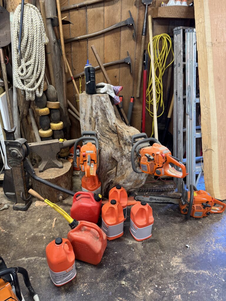 Corner filled with forestry and woodworking equipment, including chainsaws, fuel containers, an anvil, ropes, axes, ladders, and hand tools arranged around a large tree stump and wooden walls.