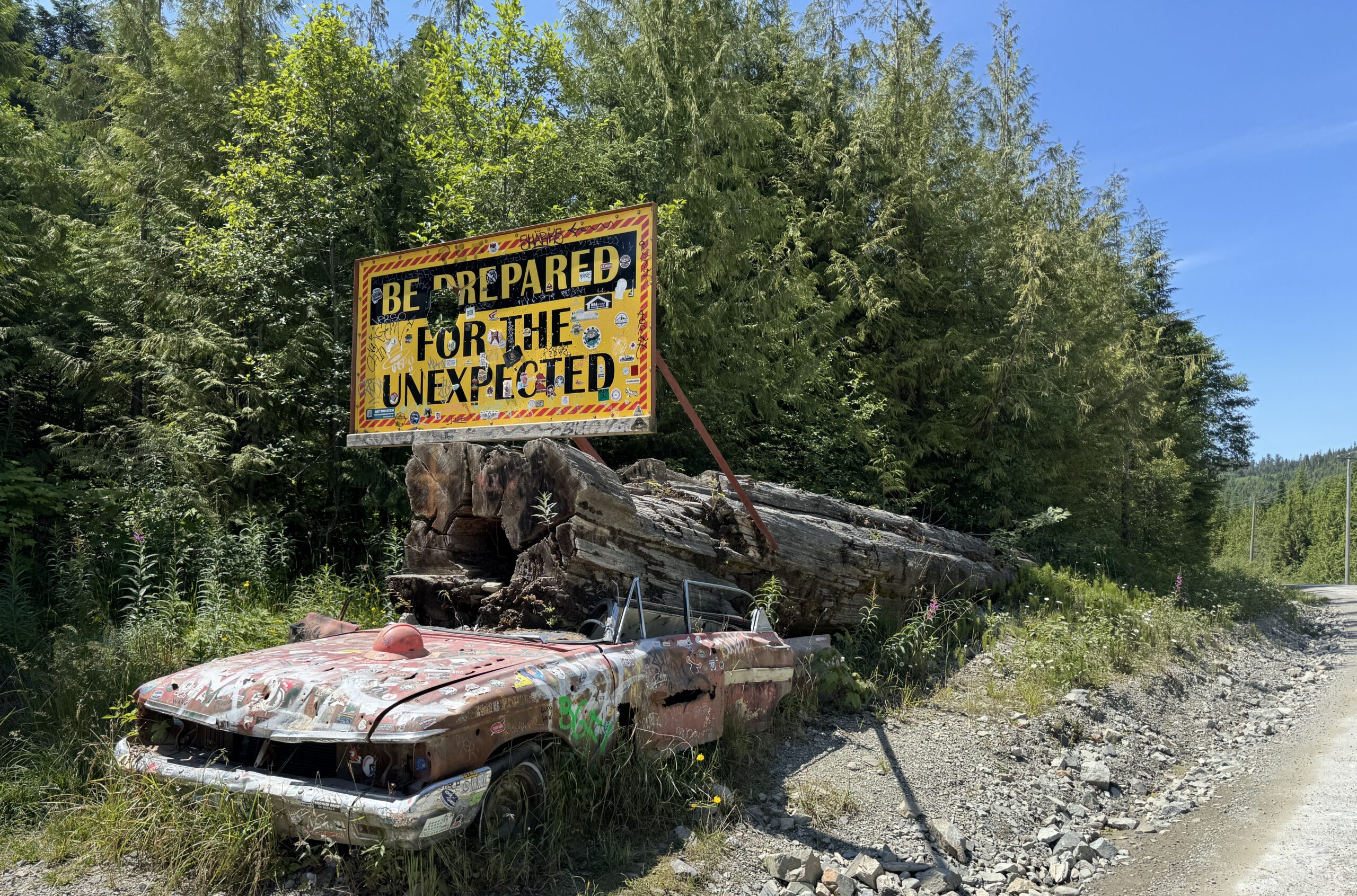 Weathered convertible covered in stickers beside a gravel forest road, with a large yellow sign mounted on a fallen log reading 'Be Prepared for the Unexpected.' Surrounded by dense evergreen trees under a clear blue sky.