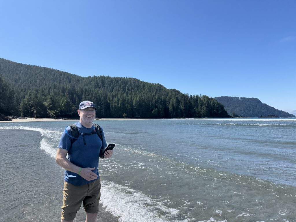 Person standing in shallow ocean water along a forested coastline, with gentle waves and tree-covered mountains in the background.