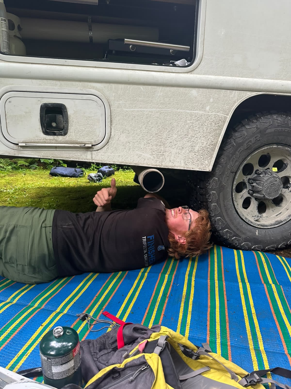Person lying on a camping mat beneath an overland camper vehicle, giving a thumbs-up while working near the exhaust and rear tire at a campsite.