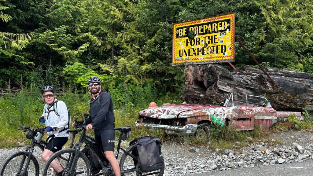 Two cyclists stop beside a weathered, graffiti-covered convertible and a large roadside sign reading “Be Prepared for the Unexpected” in a forested area.