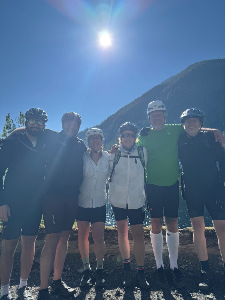 Six cyclists wearing helmets pose beside a mountain lake on a sunny day, with forested slopes and a clear blue sky in the background.